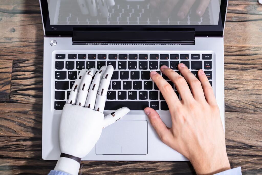 Image of one human hand and one AI hand typing at a computer.